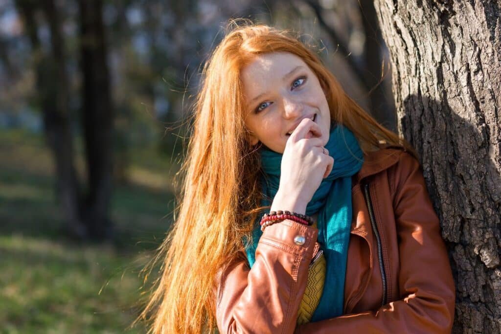Jeune femme à la chevelure rousse adossée à un arbre, main près du visage, expression timide et réservée