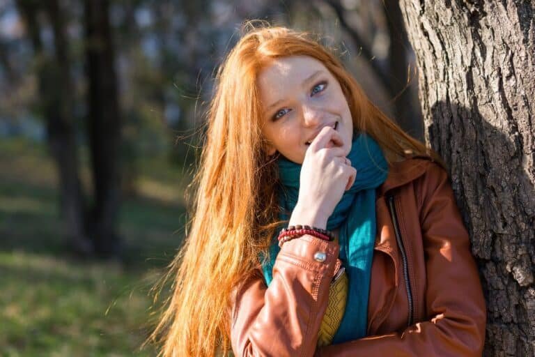 Jeune femme à la chevelure rousse adossée à un arbre, main près du visage, expression timide et réservée